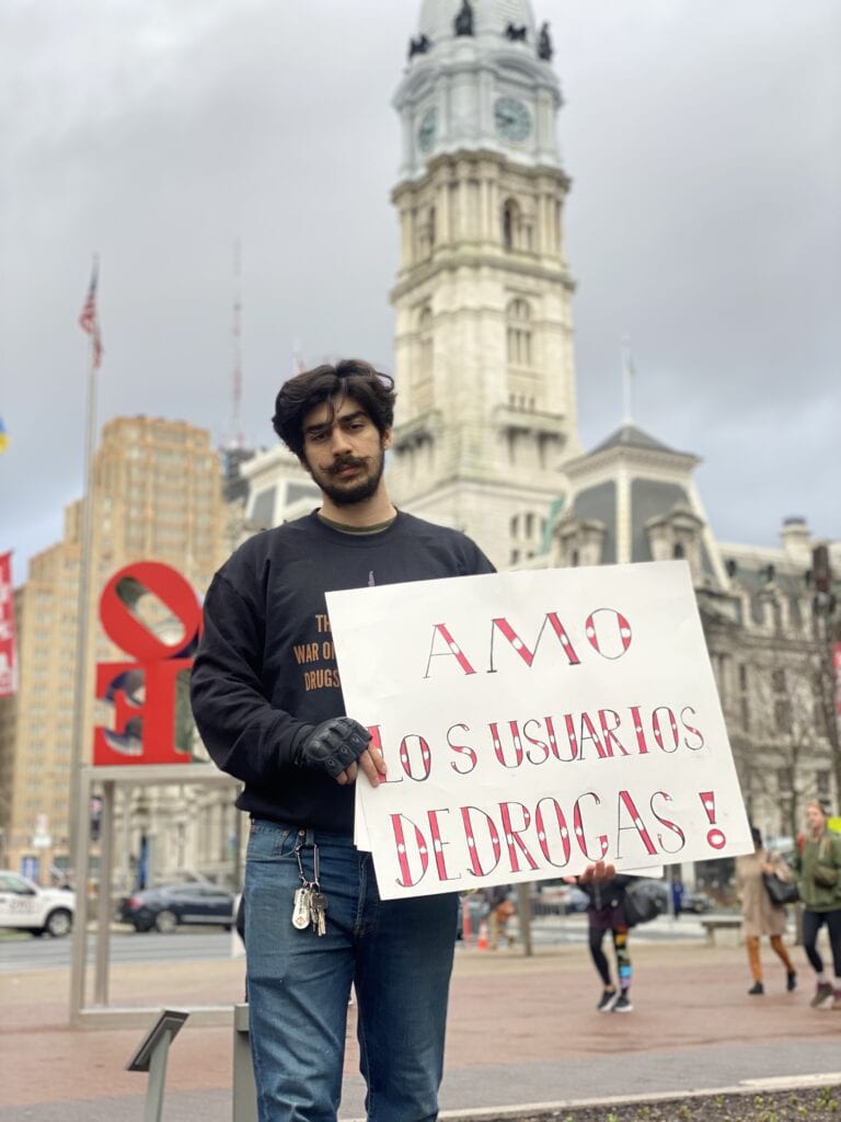 Adam Al-Asad holding a sign that says, "Amo los usuarios dedrogas!"