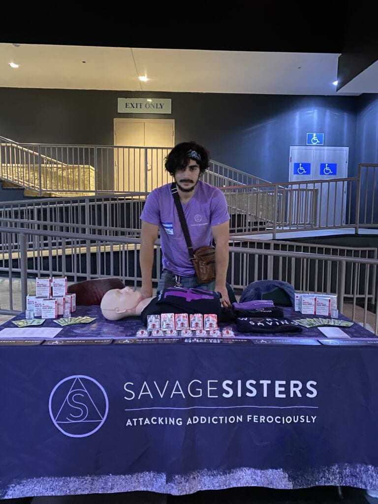 Adam Al-Asad in front of a Savage Sisters display table. The banner at the table says "Savage Sisters Attacking Addiction Ferociously"